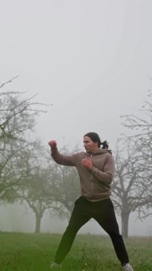 Kendini ifade etmeye adamış, etrafındaki sisli ağaçların arasında enerjik bir şekilde antrenman yapan bir insan. Dikey video. Ağır çekim. Yüksek kalite 4k görüntü