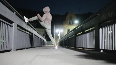 Kapüşonlu giyen yalnız bir birey, engin gece gökyüzünün altındaki boş bir köprüde enerjik bir şekilde çalışıyor. Yüksek kalite 4k görüntü