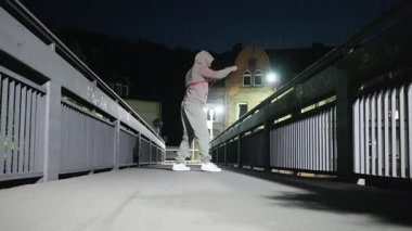 Bir insan, dayanıklılığını ve uygunluğunu vurgulamak için geceleri köprüde direnç bandı egzersizleri yapıyor. Ağır çekim. Yüksek kalite 4k görüntü