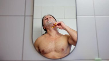 Bir adam banyo aynasının önünde tıraş oluyor ve kendi bakımının bir parçası olarak görünüşüne odaklanıyor.