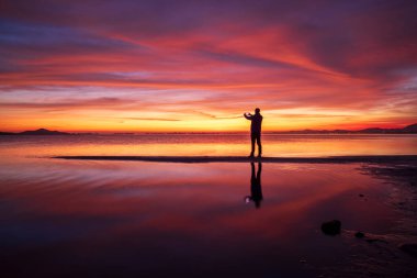 Los Urrutias sahilinde, Mar Menor, Cartagena 'da, denizin sakin sularında yansıyan bulutlarla ve cep telefonuyla güneşin doğuşunu fotoğraflayan bir adamın siluetiyle Idyllic turuncu gün doğumu.