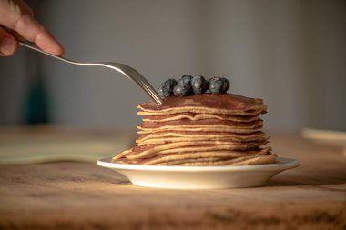 Plate of pancakes with blueberries on a wooden table and a male hand with a fork piercing one of the pancakes