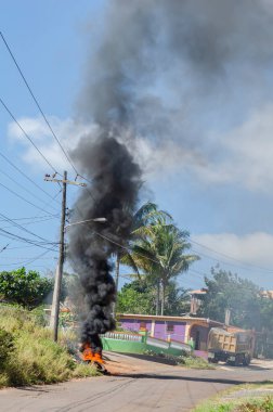 Yol kenarındaki elektrik direğinin yanındaki yanan lastiğin turuncu alevlerinin üzerinde büyük siyah ve gri duman yükseliyor..