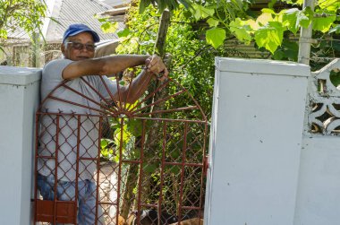 Mavili bir adam arka bahçe kapısında duruyor elleri kapalı, kahverengi metal kapının üzerinde dinleniyor ve köpekleri ayaklarının dibinde..