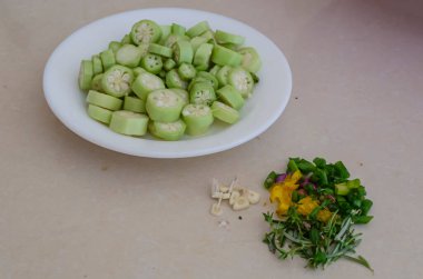 On a tile countertop there are chopped herbs beside a white plate with sliced okras. clipart