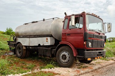 This is the side of a distributor truck with a silver insulated tank caring tar, sealcoating spray applicator, and gas cylinder, that is parked at the side of a marled road that is undergoing construction.. clipart