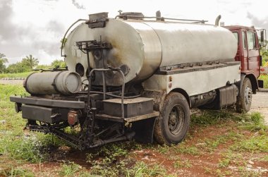 Gümüş yalıtımlı tankı olan, katran, kaplama spreyi ve gaz silindiri taşıyan bir dağıtıcı kamyon, inşaatı devam etmekte olan mermer bir yolun kenarına park edilmiştir..