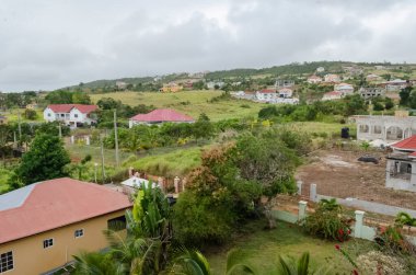 Yuvarlak tepeler, çeşitli evlerle süslenmiş, kırmızı ve beyaz çatıları olan, yemyeşil alanlar üzerine kurulmuş. Tropikal ağaçlar ve bitki örtüsü ön planda yer alırken, yeni görünümlü evler gelişmekte olan bir alanı işaret ediyor. Bulutlu gökyüzü bulutlu bir günü işaret ediyor. 