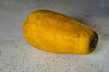 Sarı papaya beyaz benekli bir masada oturuyor. Dikdörtgen meyve, olgunluk ve tüketime hazır olma durumunu gösteren dokulu bir yüzey sergiliyor. Tablo, düz, açık renkli arkaplanı ile daha koyu benekli bir karşıtlık sağlar..