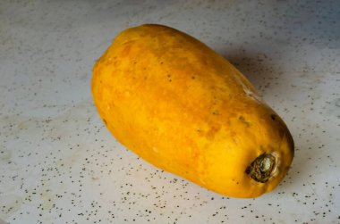 Sarı papaya beyaz benekli bir masada oturuyor. Dikdörtgen meyve, olgunluk ve tüketime hazır olma durumunu gösteren dokulu bir yüzey sergiliyor. Tablo, düz, açık renkli arkaplanı ile daha koyu benekli bir karşıtlık sağlar..