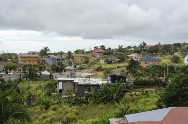Yeşillik içinde dağınık evler barındıran kırsal bir arazi. Bazı spor düz çatıları ve diğerleri sunum yaparken, evler stil ve renk olarak farklılık göstermektedir. Bölgeyi palmiye ağaçları ve yoğun bitki örtüsü sarar. Gökyüzü gri bulutlarla kaplıdır..