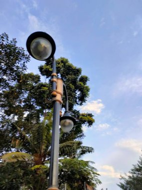 Mavi gökyüzü ve yeşil ağaçlara karşı çift yuvarlak ışıkları olan siyah sokak lambası. Kentsel, tasarım veya açık hava temalı görsel içerik için ideal.