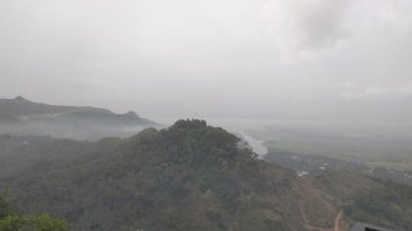 Puslu vadi manzarası, ormanlık tepeler, kıvrımlı nehir ve bulutlu gökyüzünün altındaki uzak yerleşim yerleri. Doğa, manzara veya seyahat temalı görsel içerik için ideal