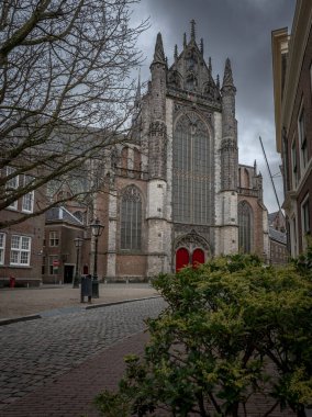 Hooghlandse  Gothic-style church, Leiden  with tall, intricate windows and red doors stands prominently at the end of a cobblestone street, surrounded by historic buildings and leafless trees under a cloudy sky clipart