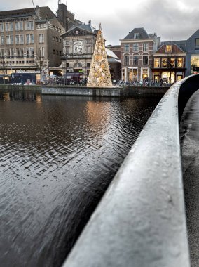 Süslü bir Noel ağacı, tarihi binalar ve dükkanlarla çevrili bir kanalın yanındaki platformda duruyor. Sahne, suyun üzerinde yansımaları ve yukarıda bulutlu bir gökyüzü olan bir köprüden izleniyor..