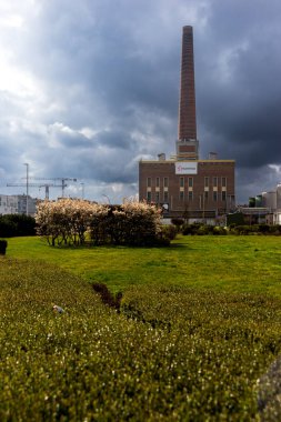 303.03.2025 Gent, Belçika, bir Luminus elektrik santralinden yükselen uzun tuğla bacası dramatik bulutlu gökyüzünün altında yeşil çimenler, budanmış çalılar ve ön planda çiçek açan beyaz ağaçlar..