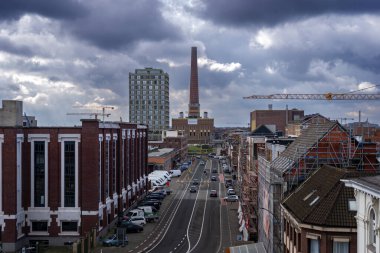 303.03.2025 Gent, Belçika, kentsel cadde. Arabaları, tadilatı devam eden binaları, inşaat vinçleri ve dramatik bulutlu gökyüzünün altında uzun bir tuğla baca bacası..