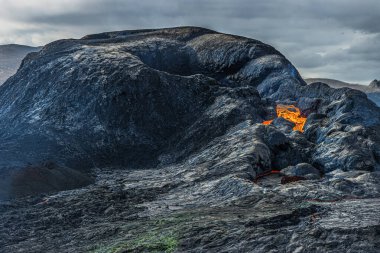 Gündüz vakti volkanik bir kraterin açılması. Lav akışına başlayan krater. Reykjanes yarımadasının İzlanda 'sındaki volkanik manzara. Kraterin etrafındaki koyu magma kayaları. Gökyüzündeki bulutlar gün ışığında