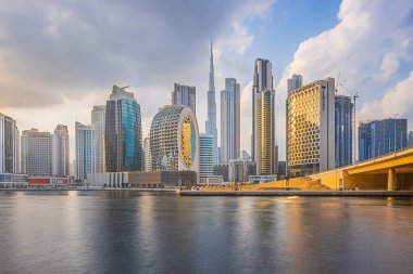 Dubai 'de günbatımı. Akşamları gökdelenler ve Burj Halife. Dubai limanında bulutlu bir gökyüzü ve liman var. Cam cepheli evler ve beton cepheli mimari.
