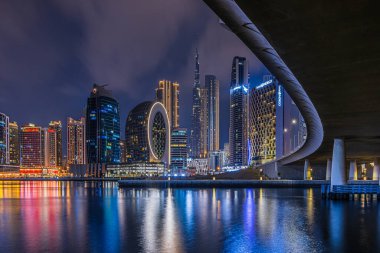 Gece Dubai silueti manzarası. Birleşik Arap Emirlikleri 'ndeki şehrin finans merkezi. Gökdelenler mavi saatte bir köprünün kıvrımlı rotasıyla aydınlandı. Su yüzeyindeki yansımalar