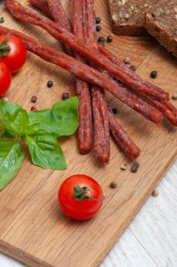 Thin hunting sausages on a wooden board decorated with tomatoes and basil top view