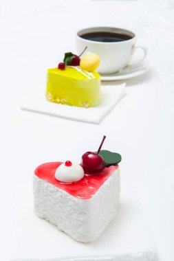 Bir fincan kahve, kalp şeklinde bir kek, kırmızı jöleli, açık arkaplanda bir tabakta.