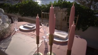 Şamdanlardaki pembe mumlar deniz kıyısındaki terasta serilmiş bir masanın üzerinde duruyor. Yüksek kaliteli FullHD görüntüler