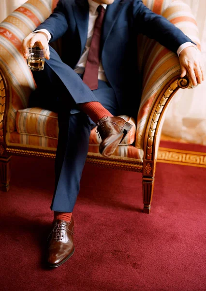 Legs of a man in blue pants, red socks and brown shoes sitting in the chair holding glass in one hand. High quality photo