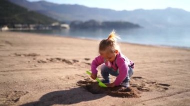 Küçük kız kumsalda kumsalda bir çukur kazıyor, yere çömelmiş bir oyuncak kürekle. Yüksek kalite 4k görüntü