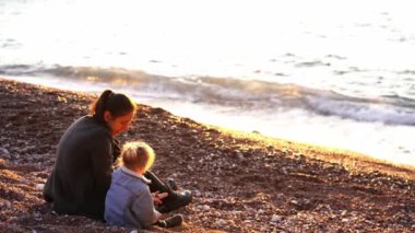Mom and little daughter are sitting on a pebble beach and sorting through the pebbles. High quality 4k footage