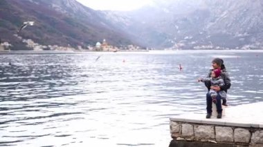 Mom with a small child in her arms sits on a pier by the sea and looks at the flying seagulls. High quality 4k footage