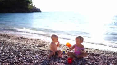 Küçük bir oğlan ve kız, deniz kenarındaki çakıl taşlı bir plajda plastik kovalarla oturuyorlar. Yüksek kalite 4k görüntü