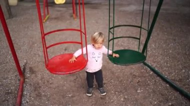 Little girl swings a swing while standing next to them on the playground. High quality 4k footage