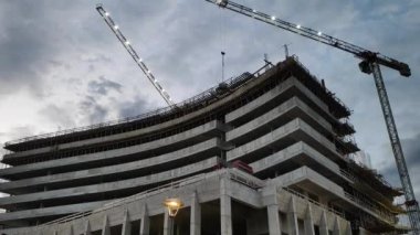 Illuminated tower cranes work at the construction site of a multi-storey building against a gloomy sky. High quality FullHD footage