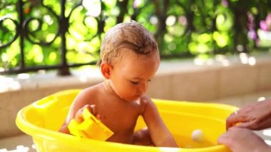 Mom splashes water on a little girl sitting in a bowl on the balcony with toys in her hands. High quality 4k footage