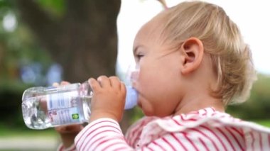 Little girl drinking water from a bottle standing in the park. High quality FullHD footage