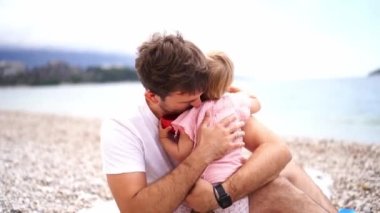 Dad hugs a little girl and sits her on his lap on the beach by the sea. High quality 4k footage