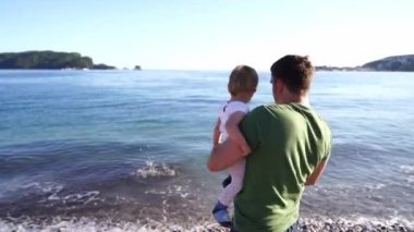 Dad with a little girl in his arms throws pebbles into the sea. High quality 4k footage