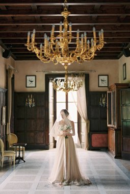 Bride in a veil with a bouquet of flowers stands against the background of a window in the room. High quality photo