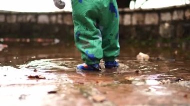 Small child in overalls and rubber boots walks through a puddle in the rain. High quality 4k footage