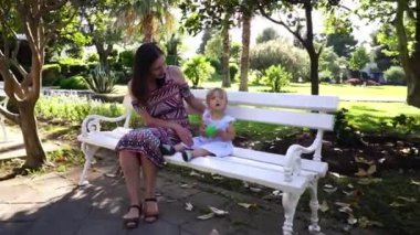 Mom and little girl are sitting on a white bench in a green park. High quality FullHD footage