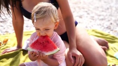 Little girl eating a piece of watermelon while sitting on a blanket on the beach next to her mother. High quality 4k footage