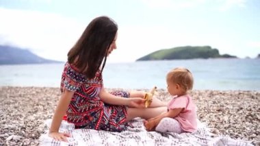 Mom gives a little girl a banana while sitting on a blanket on the beach by the sea. High quality 4k footage