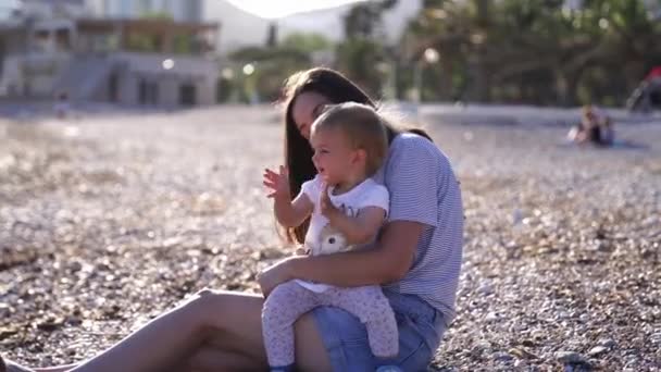 小女孩坐在妈妈的腿上 坐在一块铺着鹅卵石的海滩上 一边拍手 高质量的4K镜头 — 图库视频影像