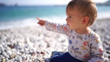 Little girl sits on a pebble beach, points to the surf and babbles. High quality 4k footage