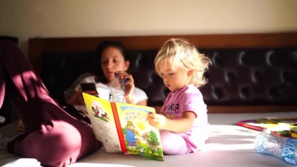 Маленька Дівчинка Сидить Книгою Ліжку Поруч Матір Лежачи Смартфоном Кадри — стокове відео
