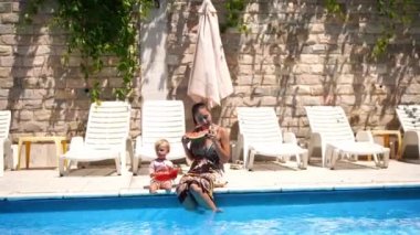 Mom with a little girl eat pieces of watermelon, sitting near the pool and putting their feet in the water. High quality 4k footage