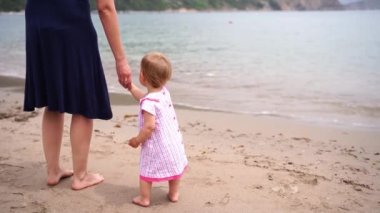 Little girl with her mom by the hand stands on the beach and looks at the sea surf. High quality 4k footage