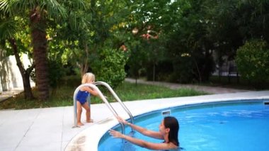 Little girl tramples her feet on the shore of the pool near her mother in the water. High quality 4k footage