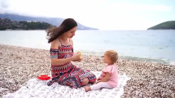 Мама Очищає Банан Маленької Дівчинки Сидячи Ковдрі Пляжі Біля Моря — стокове відео